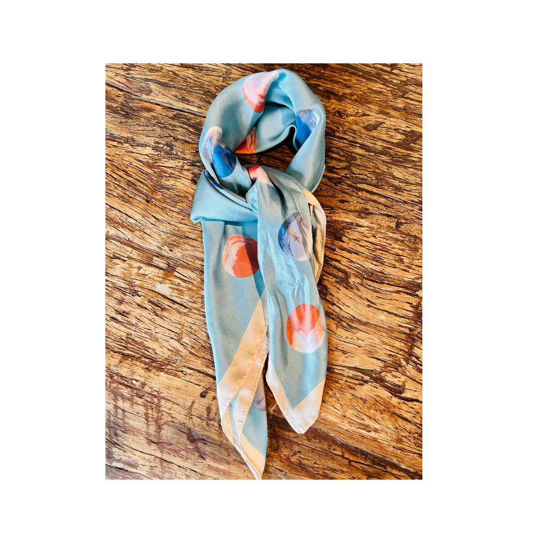 A light blue silk scarf with a colorful abstract print, draped on a rustic wooden surface.
