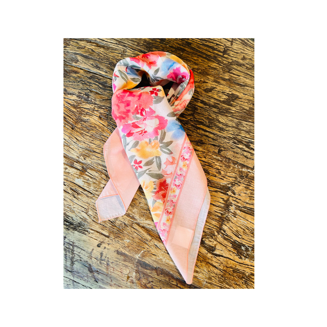 A pastel pink silk scarf with a vibrant floral print, styled in a loop on a rustic wooden surface.
