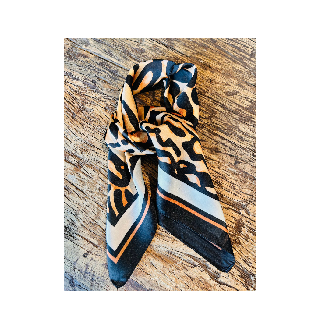 A patterned silk scarf with black, beige, and orange abstract designs and striped accents, styled on a rustic wooden surface.