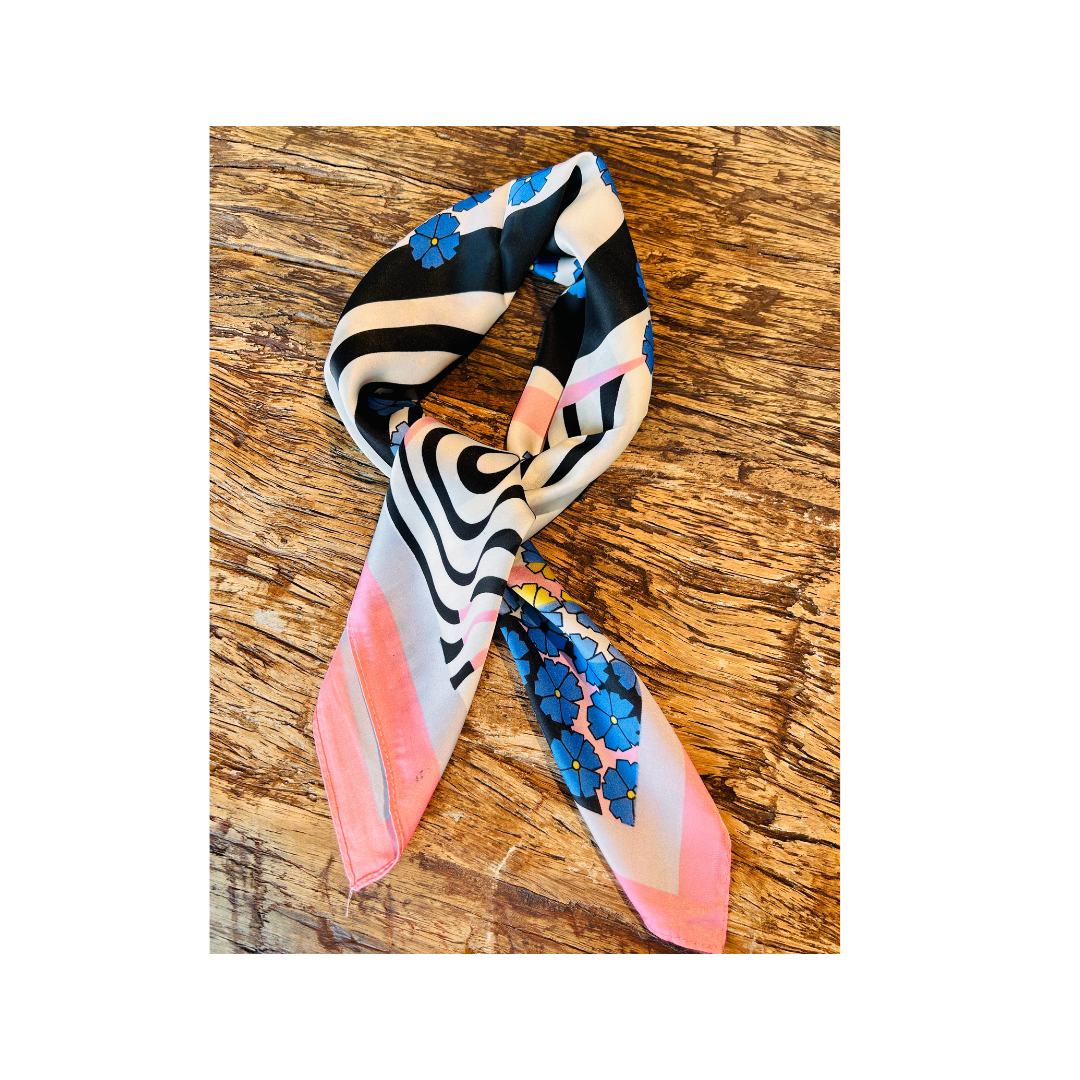 A silk scarf with a multicolor abstract pattern featuring black, white, pink, blue, and yellow, styled in a knot on a rustic wooden surface.