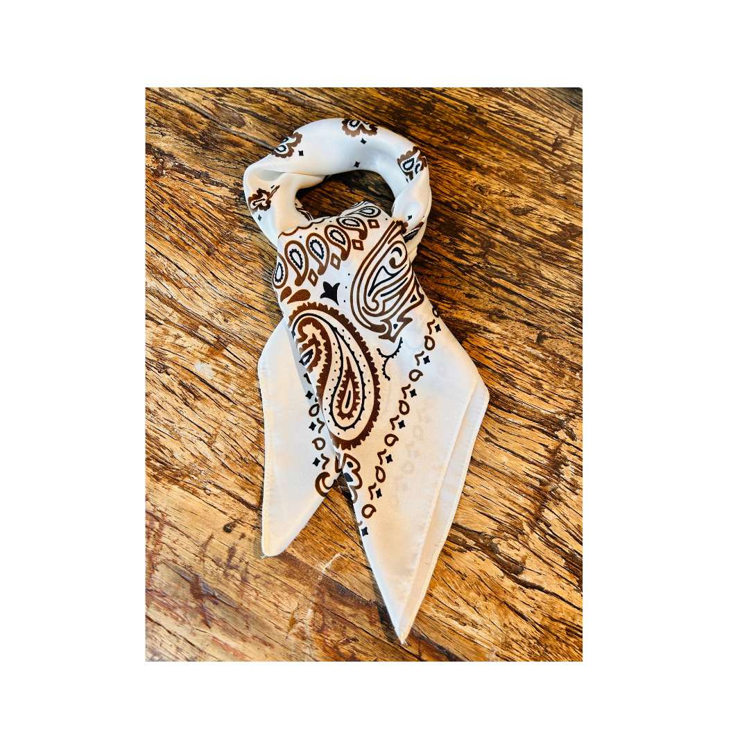 A white bandana scarf with a brown paisley pattern, folded and displayed on a rustic wooden surface.