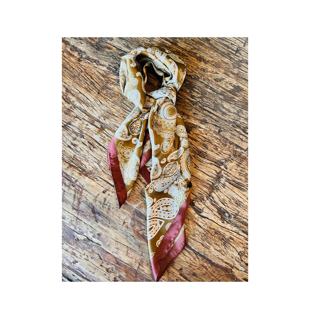 A patterned silk scarf with a cream and brown paisley design and deep burgundy ends, tied in a knot and displayed on a rustic wooden surface.