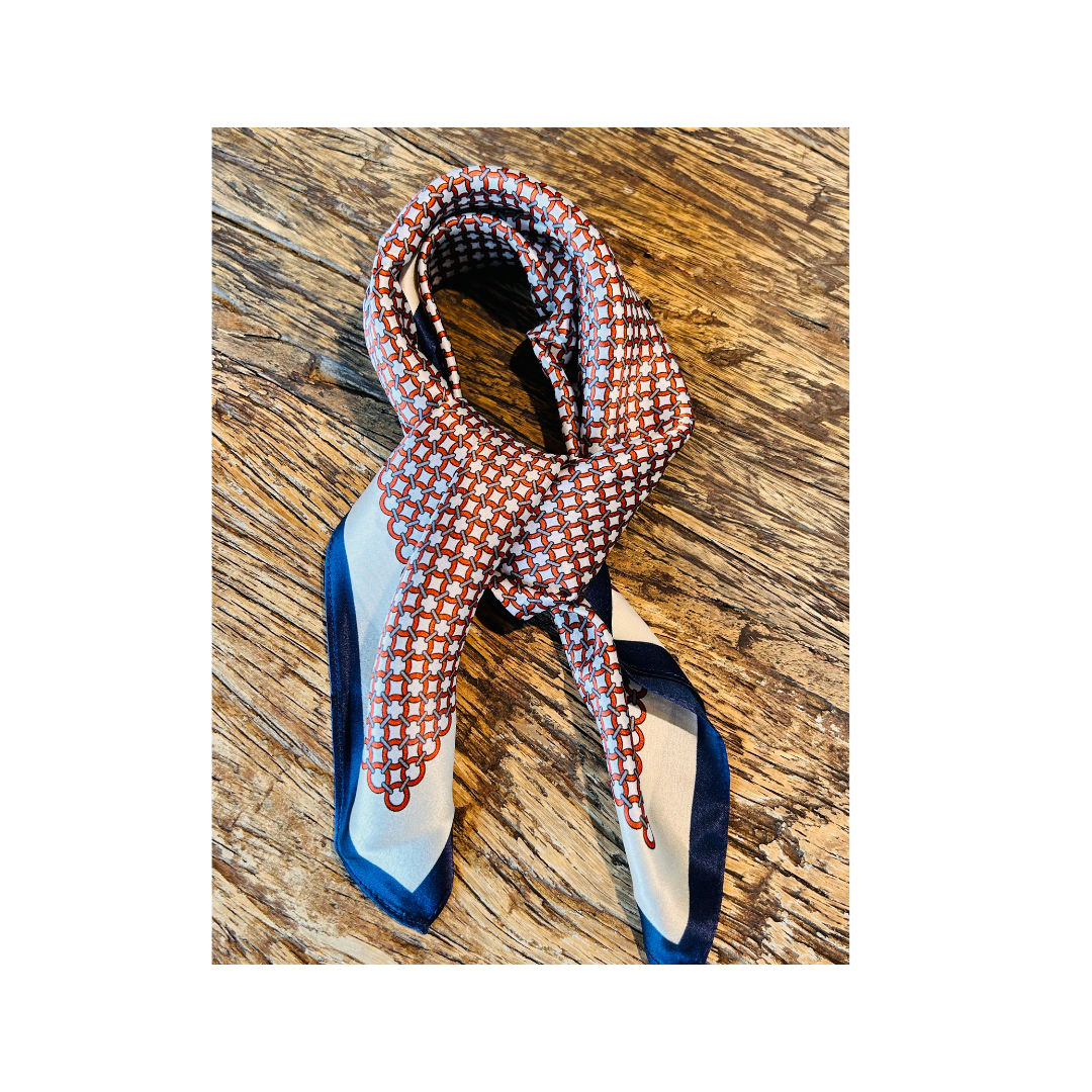 A patterned silk scarf with a geometric design in orange, white, and blue, tied in a knot and displayed on a rustic wooden surface.